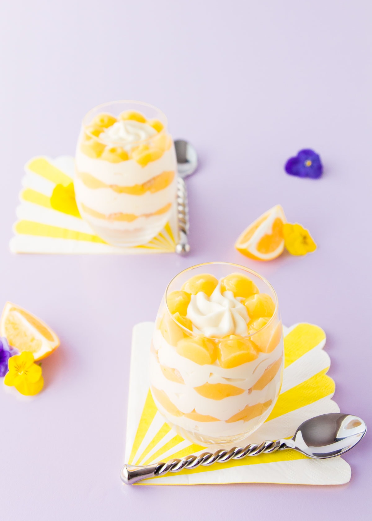 Two lemon cheesecake cups, on yellow and white striped napkins, on a pale purple background. 