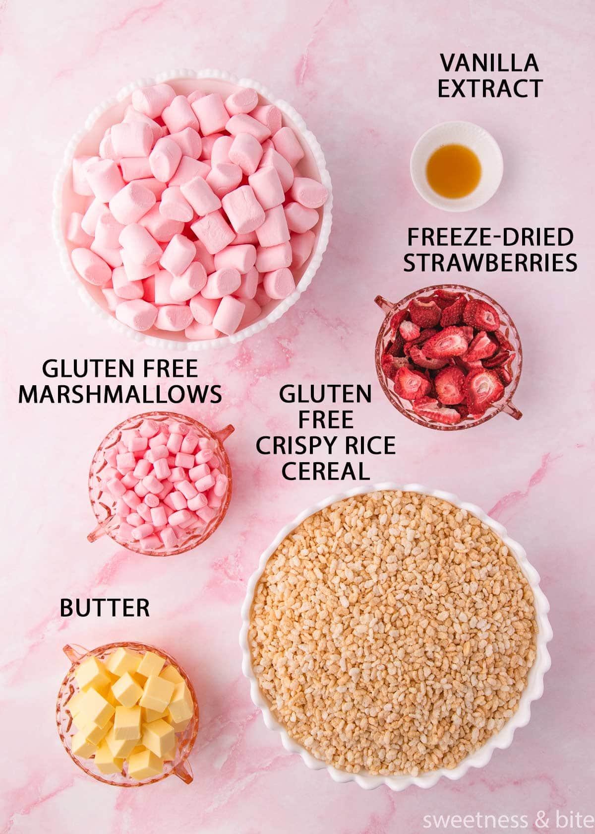 The ingredients in white bowls on a pink marble background - marshmallows, rice krispies, butter, vanilla and freeze-dried strawberries. 