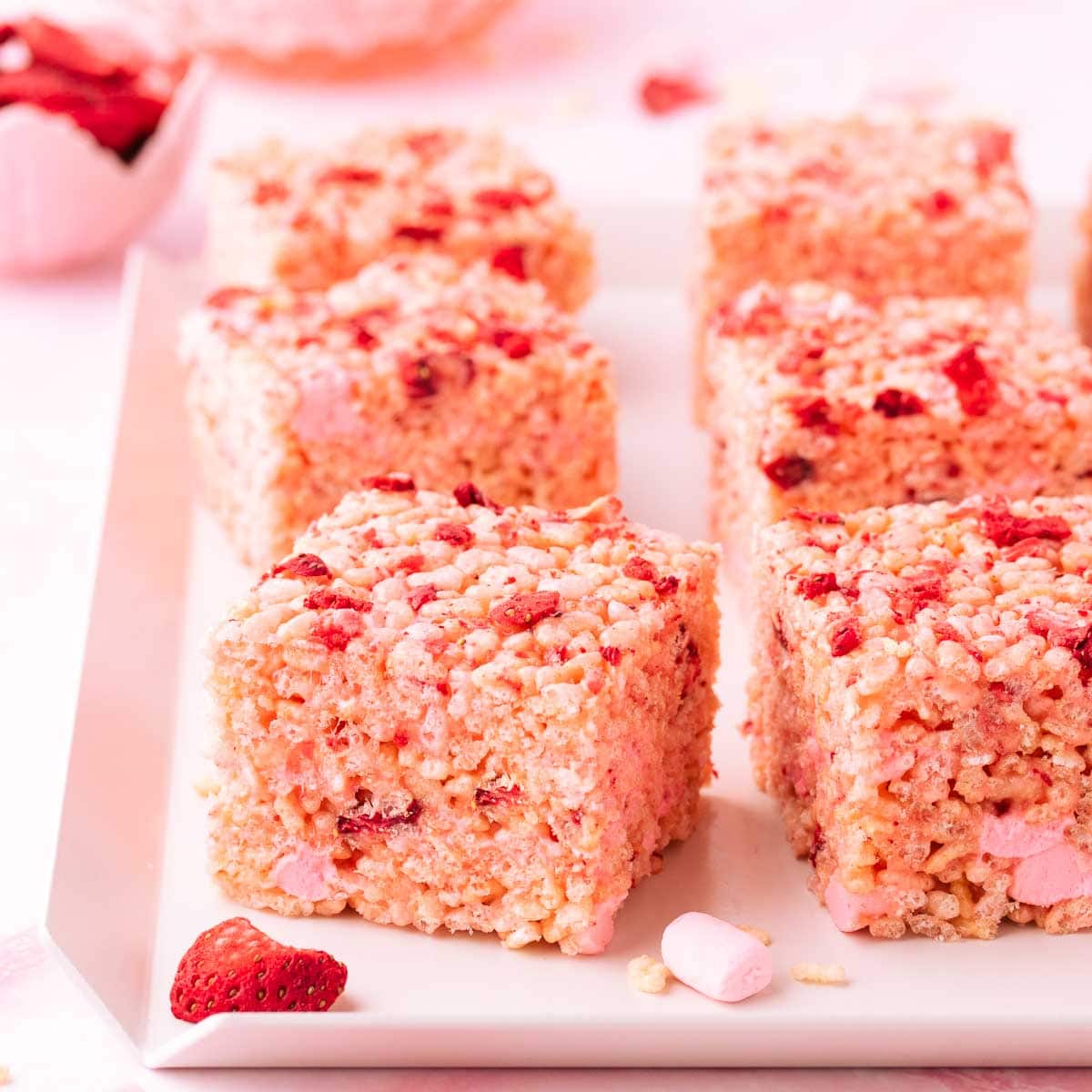 Six square strawberry rice krispie treats on a white tray with scattered pieces of freeze-dried strawberry.