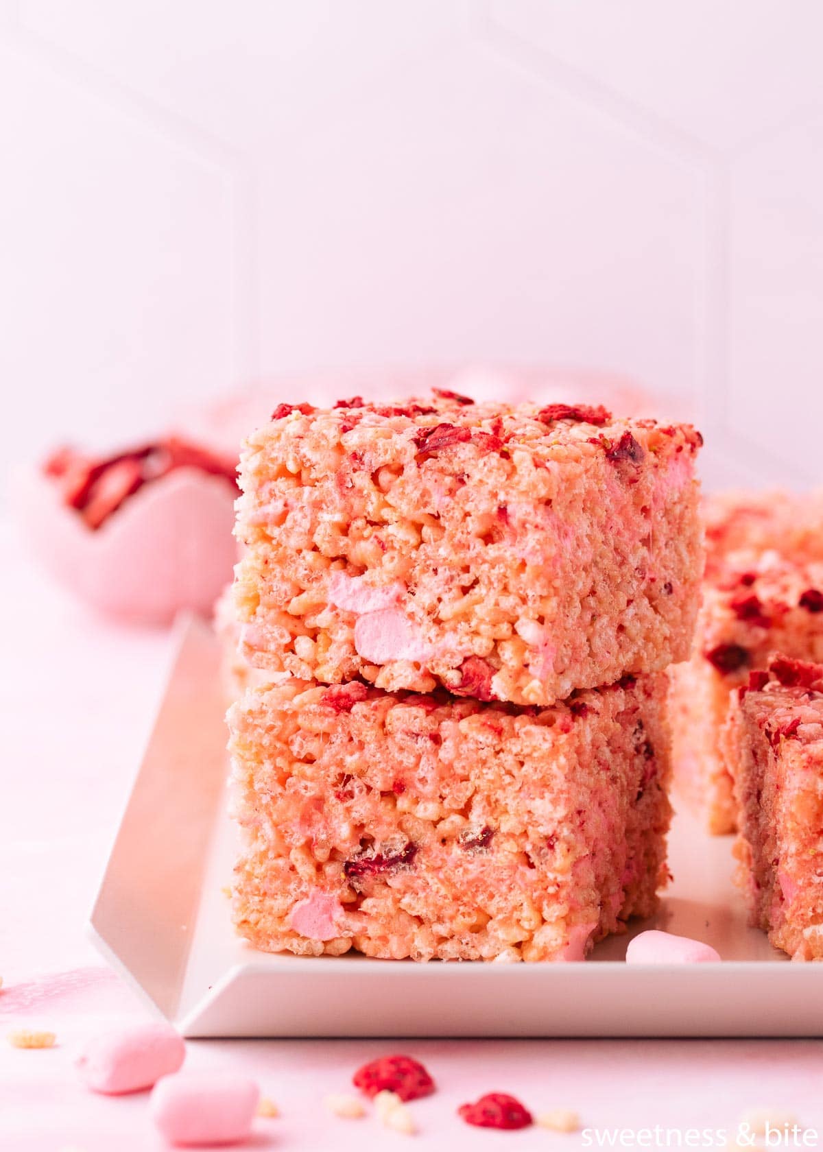 Two stacked strawberry rice krispie treats on a white tray with a small pink bowl of freeze-dried-strawberries in the background. 