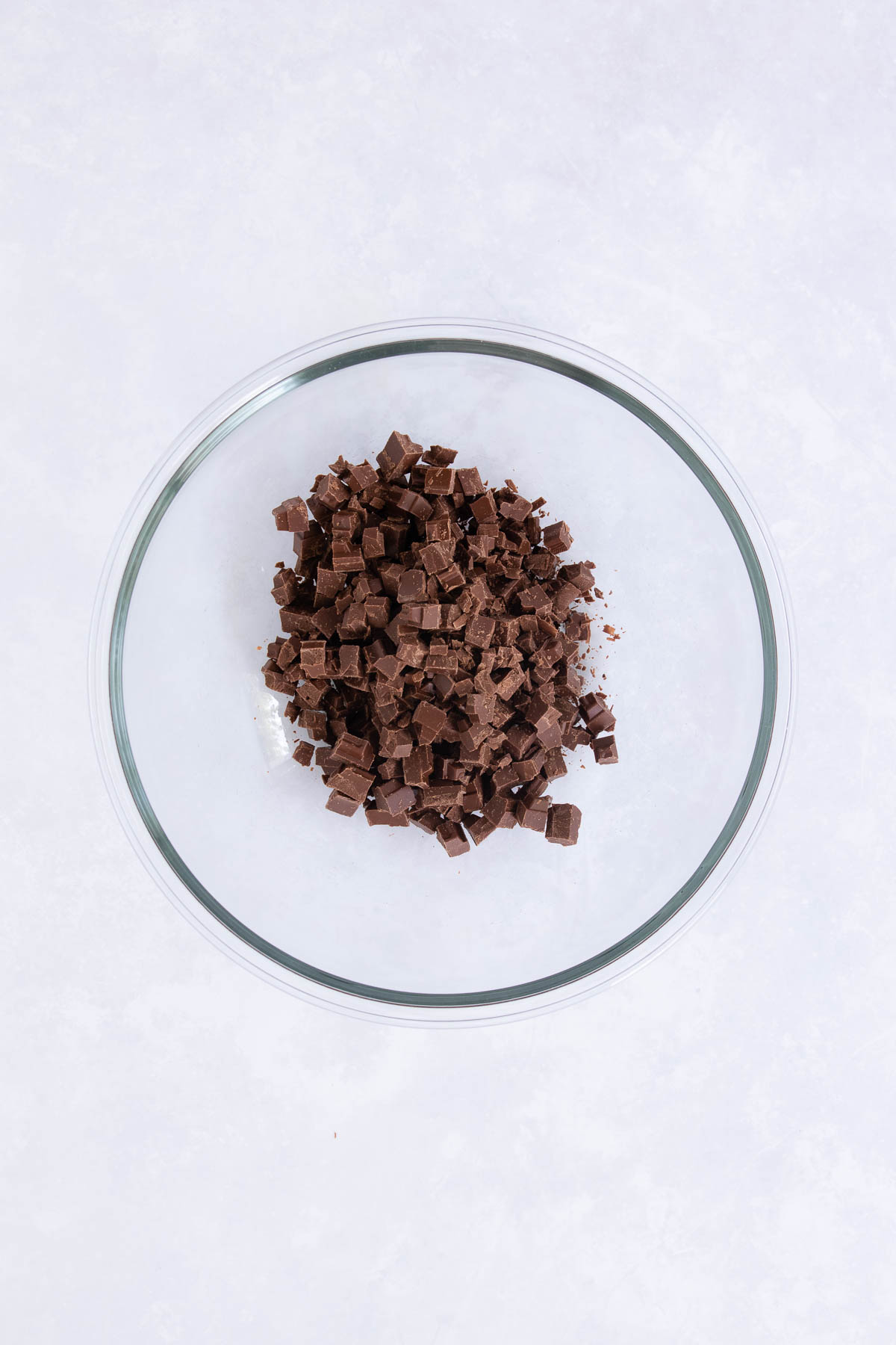 Chopped milk chocolate in a large glass mixing bowl, on a grey marble background.