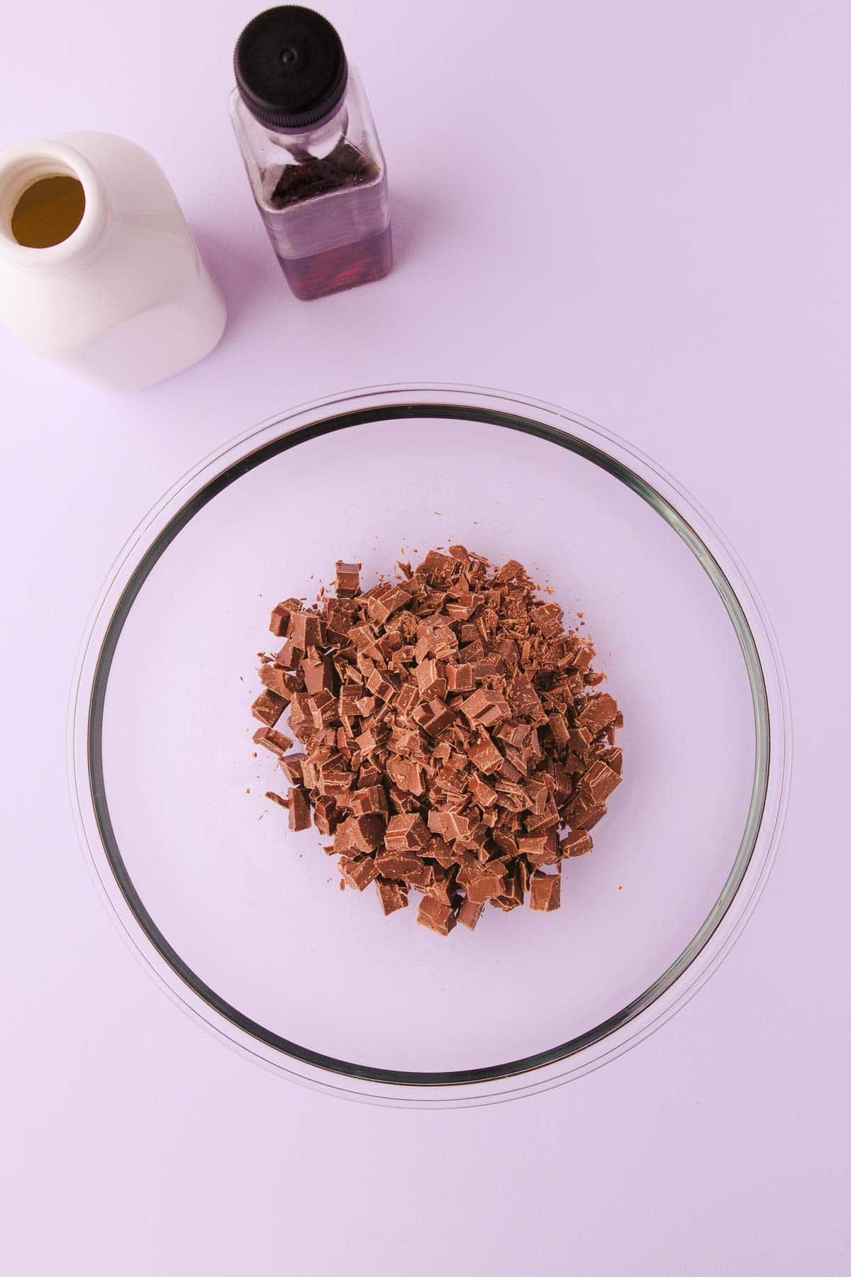 Chopped milk chocolate in a large glass bowl on a pale purple background.