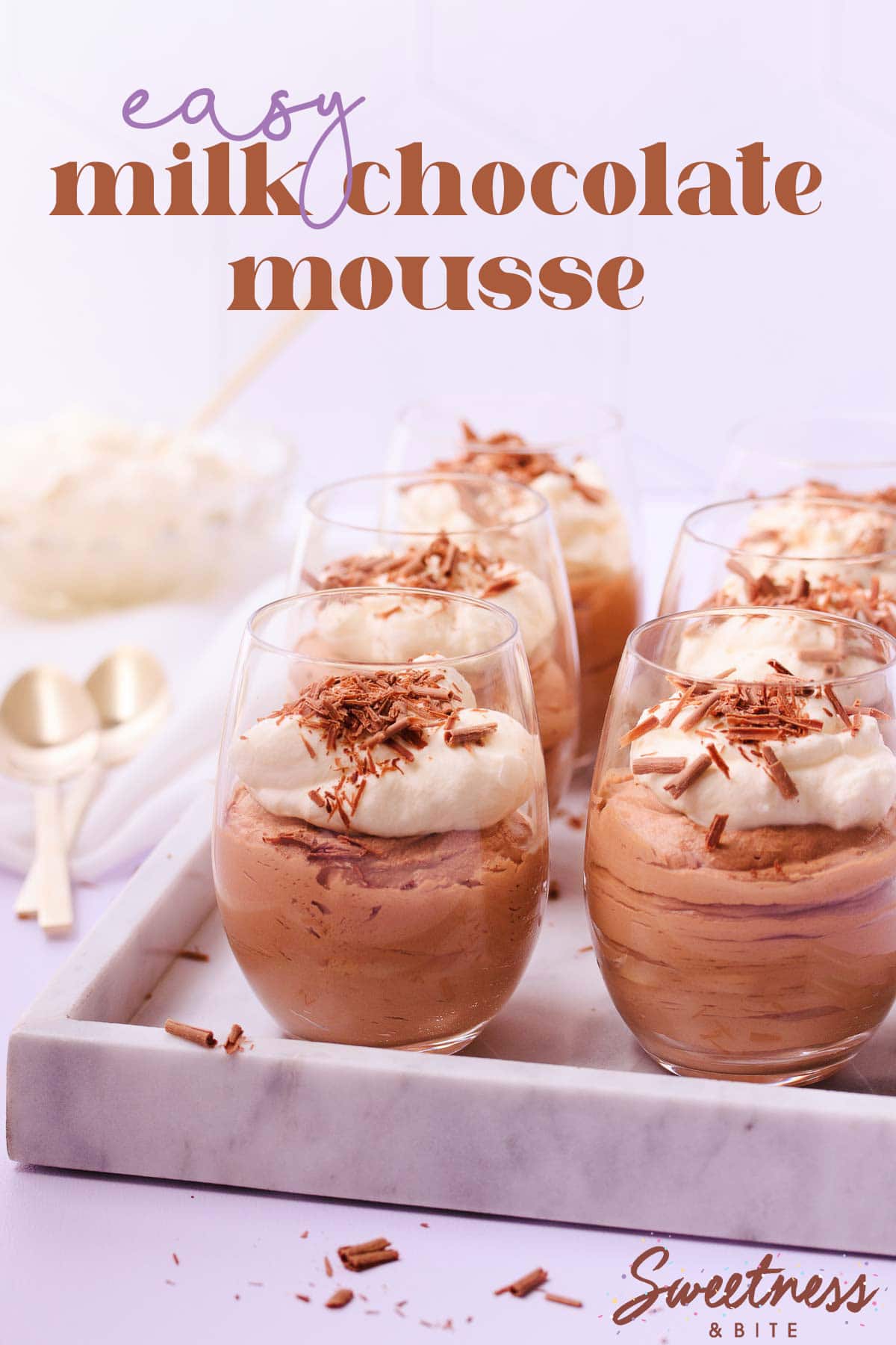 Six stemless wine glasses filled with milk chocolate mousse and topped with whipped cream and chocolate shavings, on a marble tray, with gold teaspoons and a glass bowl of whipped cream.