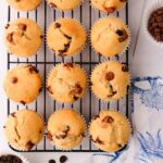 12 gluten free chocolate chip muffins in rows on a black metal cooling rack, with a blue and white napkin and a scattered chocolate chips.