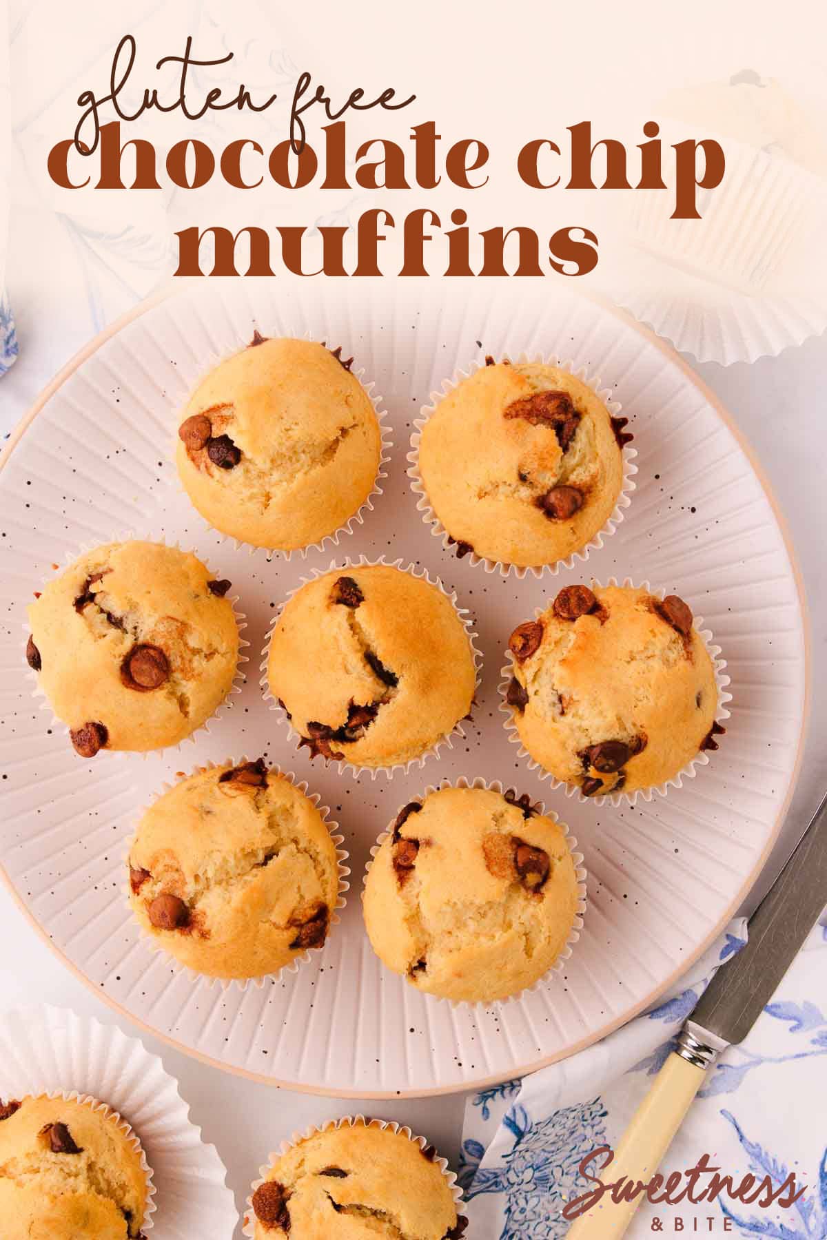 7 gluten free chocolate chip muffins arranged in a circle on a white plate with black speckles, with a blue and white napkin and a bone-handled knife.