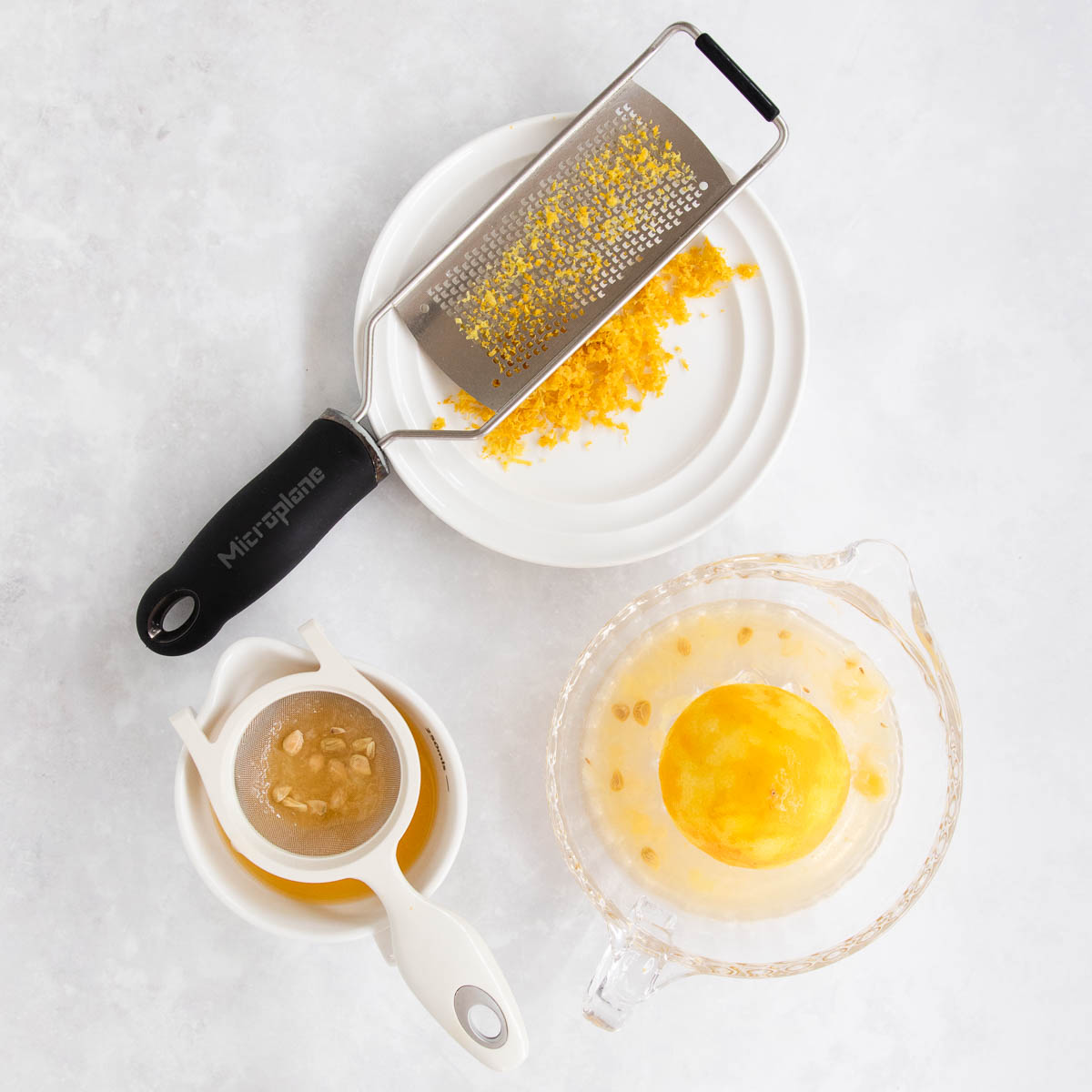 Lemon zest on a white plate with a microplane grater, and a glass lemon juicer with a juiced lemon, on a grey marble background.