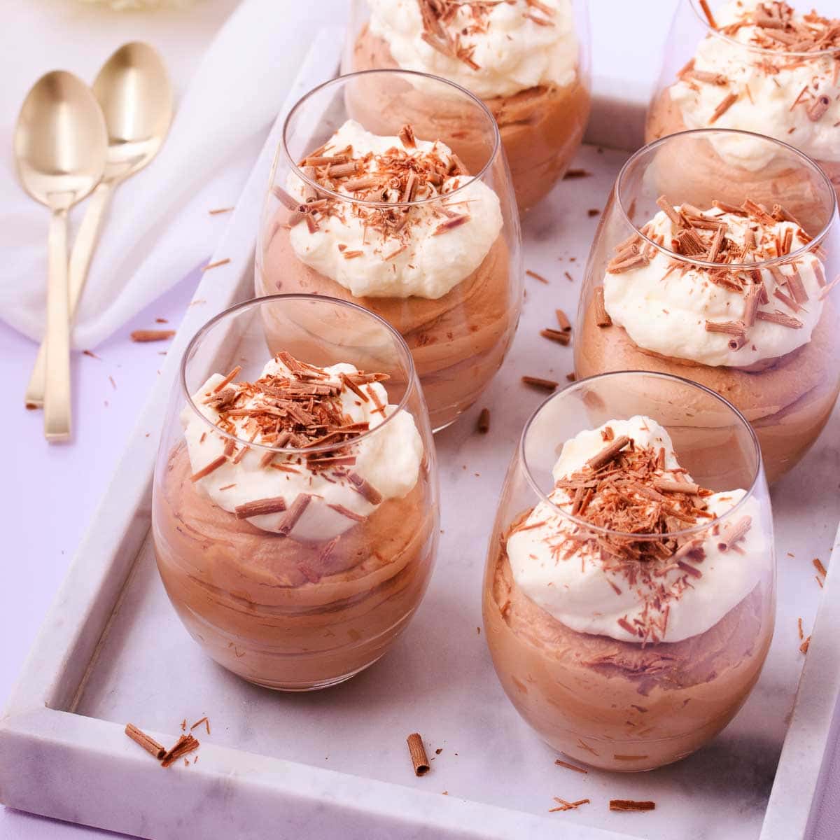 Six stemless wine glasses filled with milk chocolate mousse and topped with whipped cream and chocolate shavings, on a marble tray, with gold teaspoons.