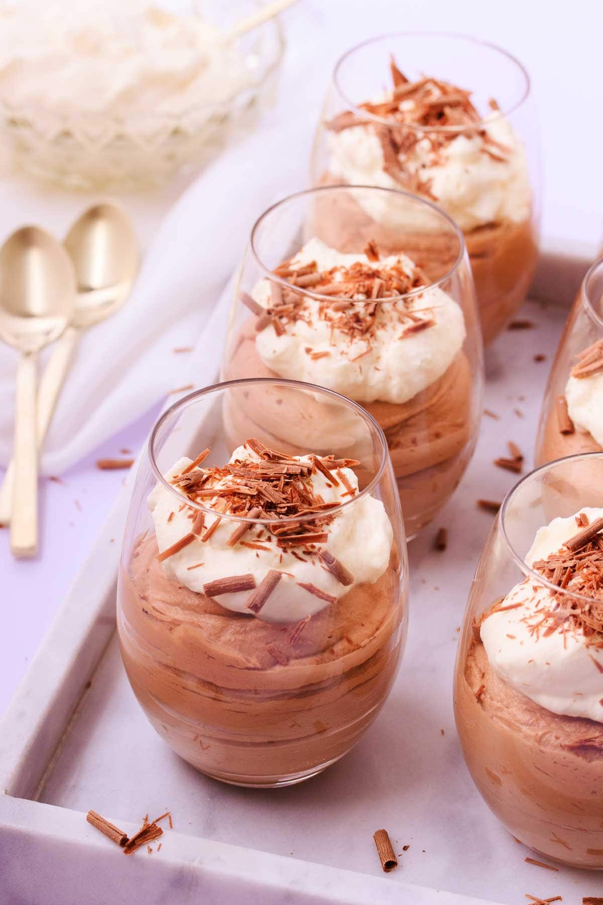 Six stemless wine glasses filled with milk chocolate mousse and topped with whipped cream and chocolate shavings, on an offset marble tray, with gold teaspoons.