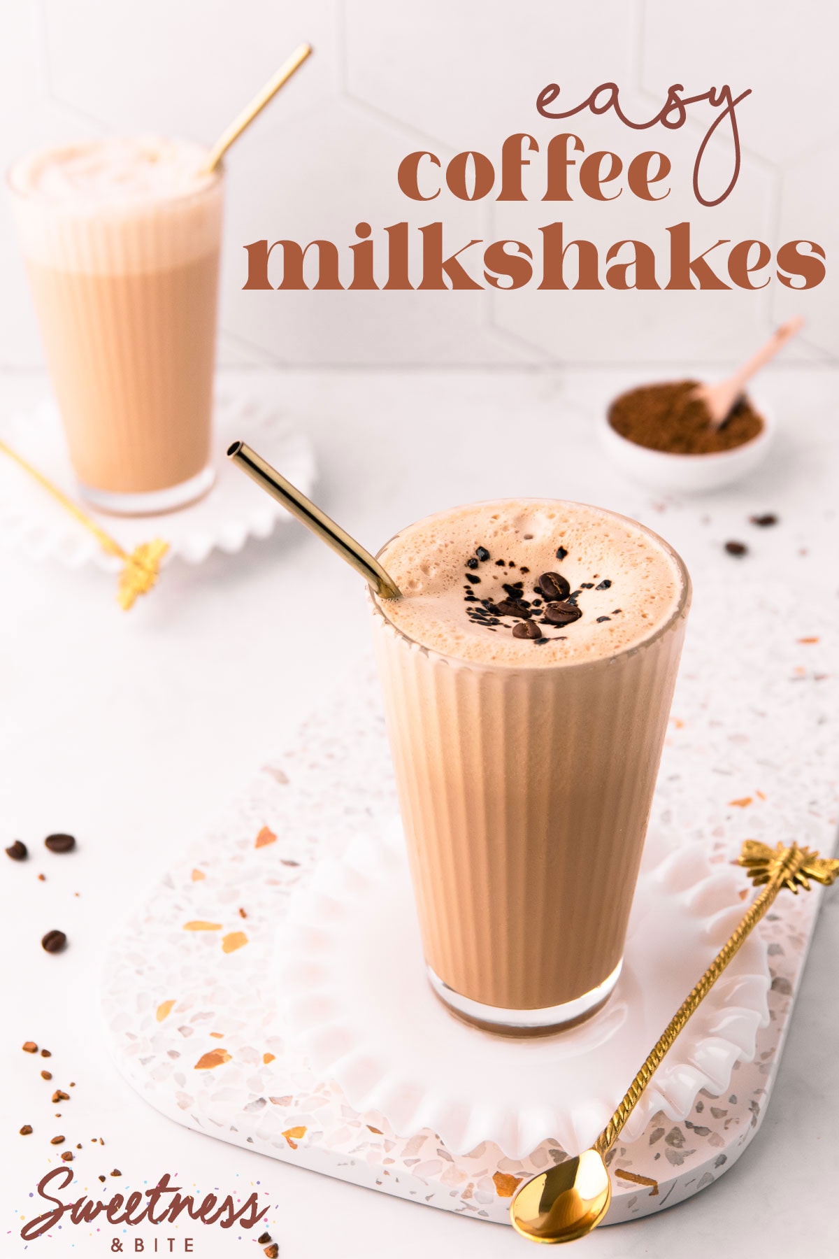 Two coffee milkshakes in tall glasses with gold straws, on a terrazzo board, with a small bowl of coffee granules.
