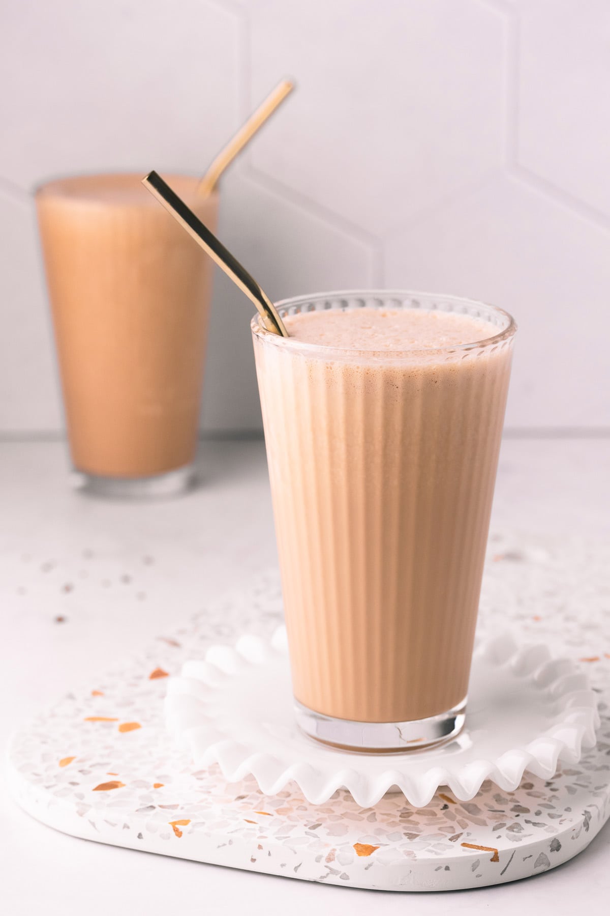 A freshly poured coffee milkshake, with a gold metal straw, on a grey tiled background.