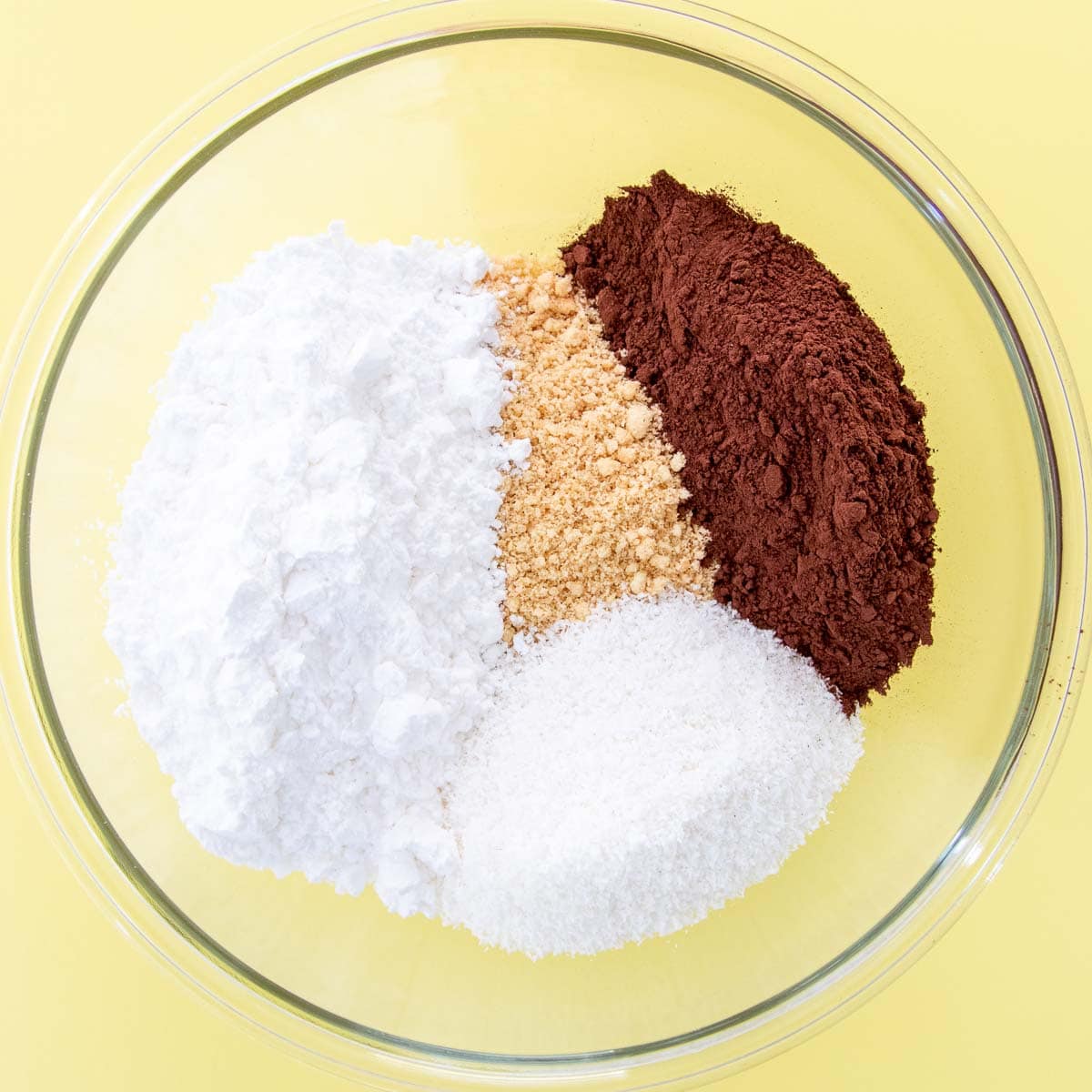 The crushed biscuits, cocoa powder, coconut and powdered sugar in a large glass bowl on a yellow background.