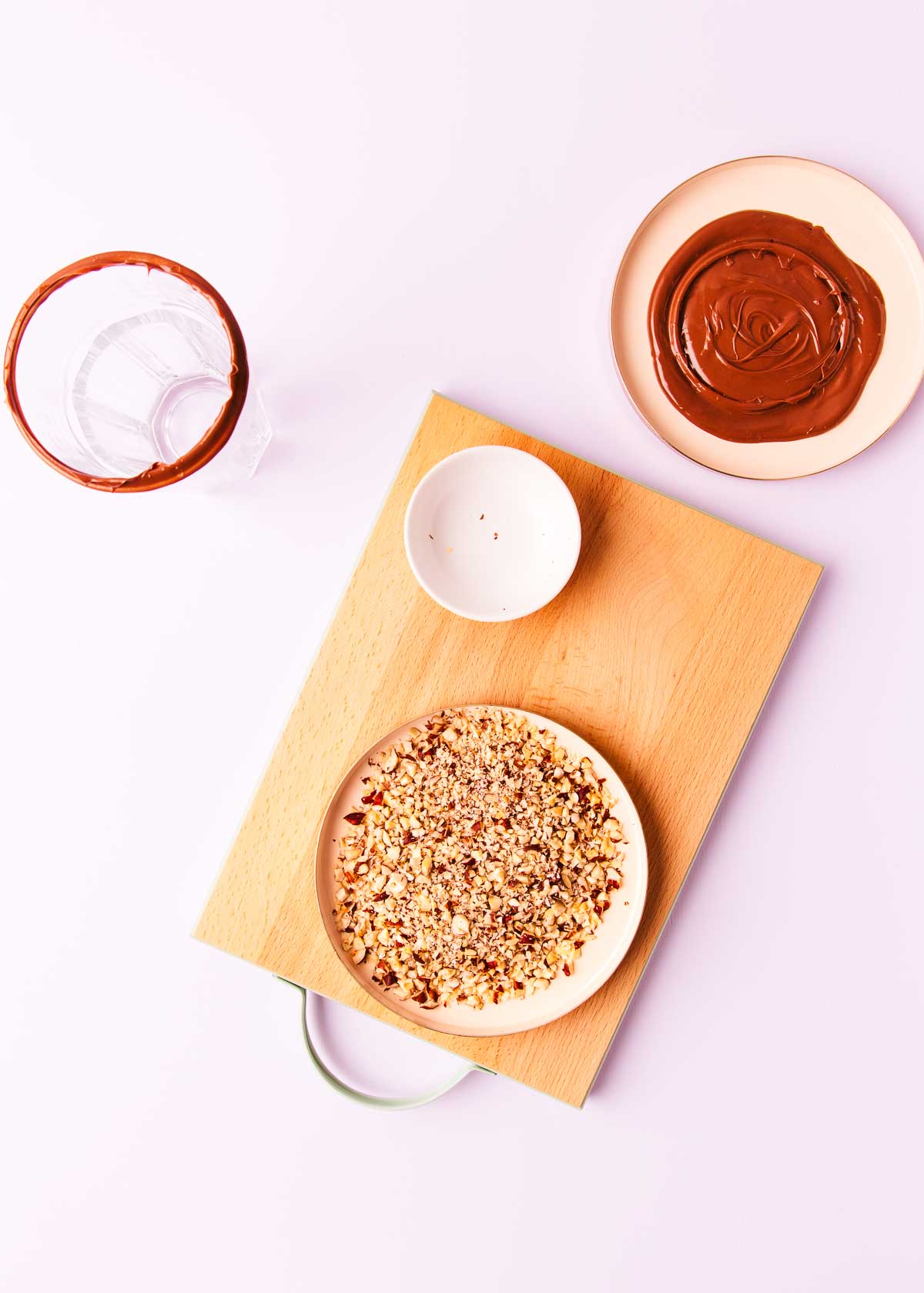 Chopped hazelnuts spread in a thin layer on a pink plate, with a Nutella-dipped glass, on a pale purple background.