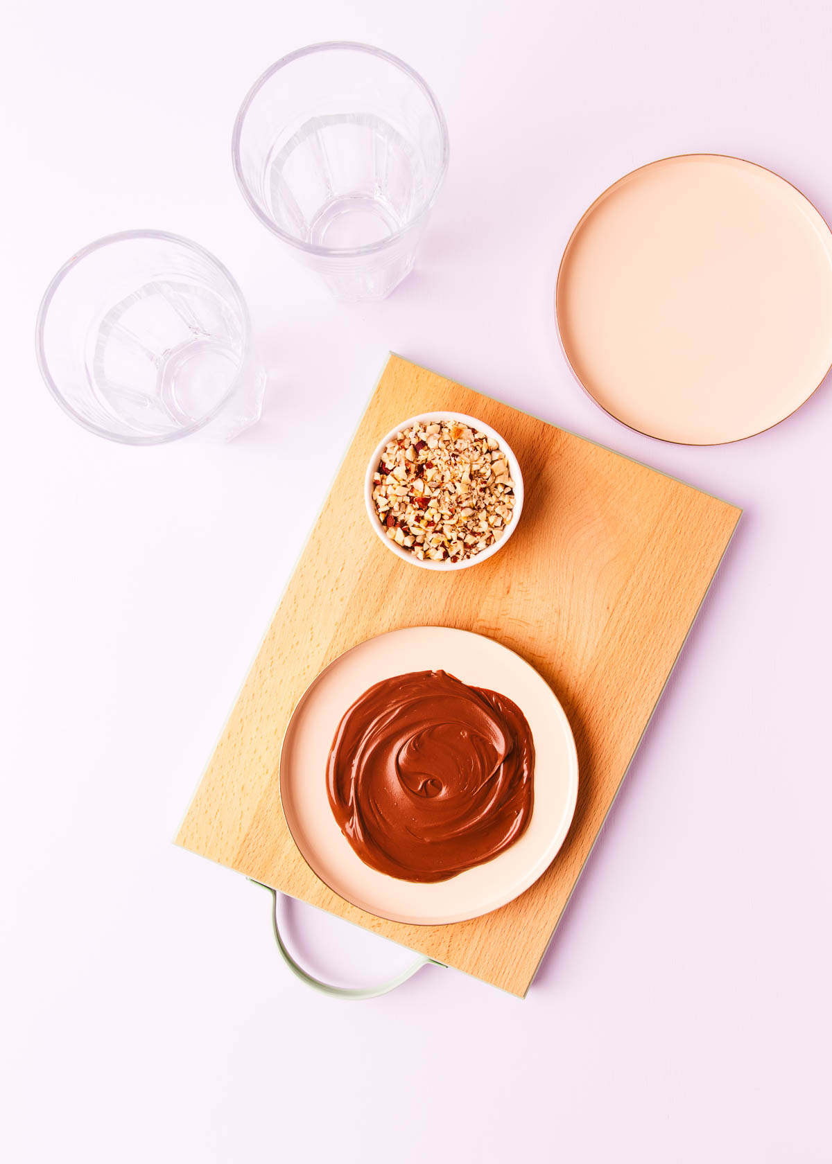 Softened Nutella spread onto a flat pink plate, with two tall empty glasses, on a pale purple background.