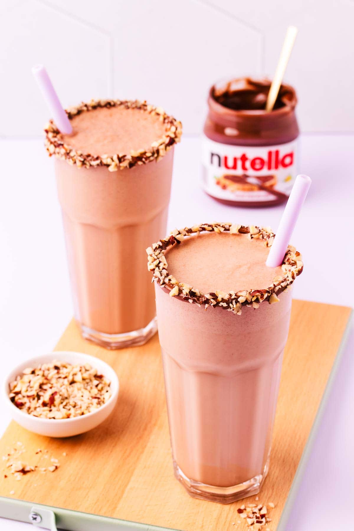 Two milkshakes with a jar of Nutella and a small bowl of chopped hazelnuts. 