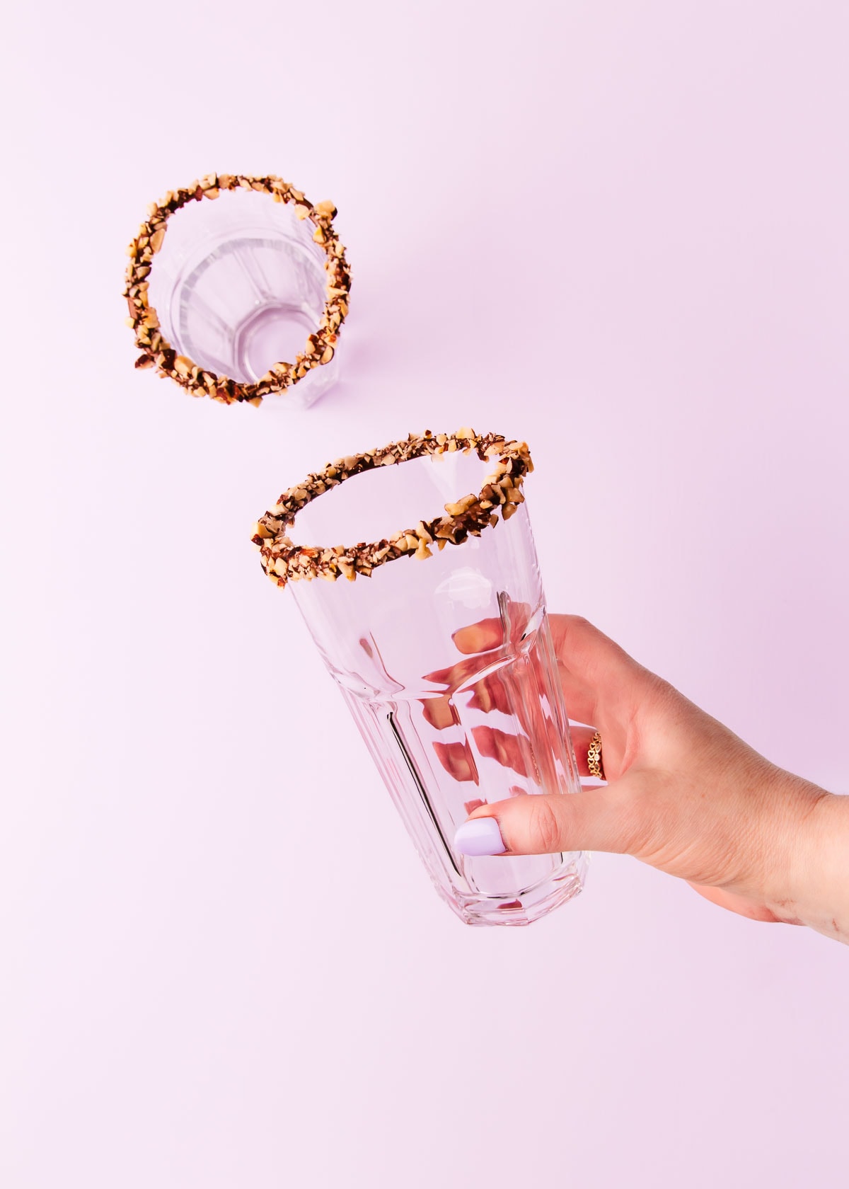 A hand holding a glass with a Nutella dipped rim covered in chopped hazelnuts.