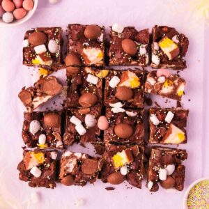16 square pieces of Easter egg rocky road on a piece of crinkled baking paper, on a light purple background, with mini Easter eggs and pastel sprinkles scattered around.