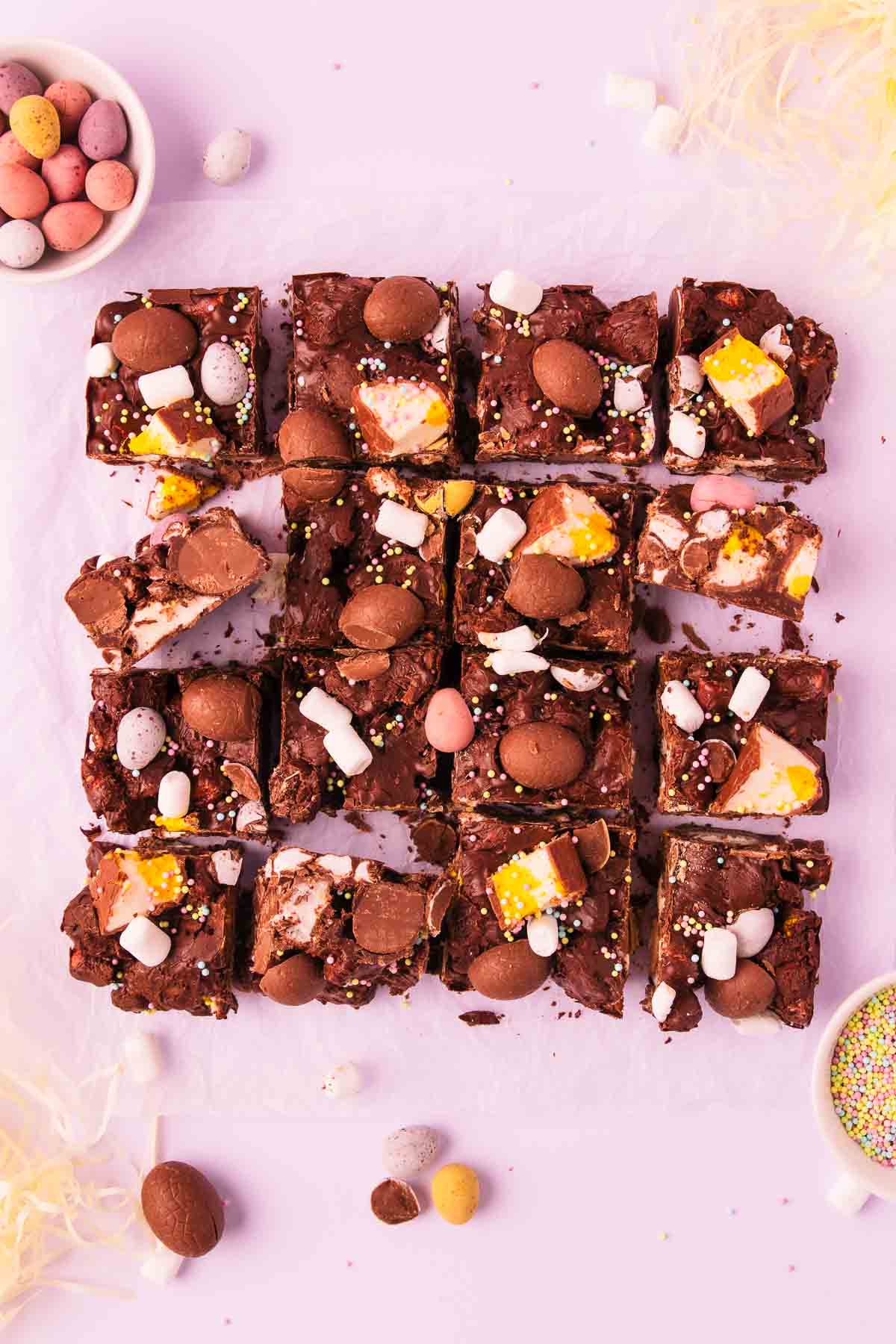 16 square pieces of rocky road on a piece of crinkled baking paper, on a light purple background, with mini Easter eggs and pastel sprinkles scattered around.