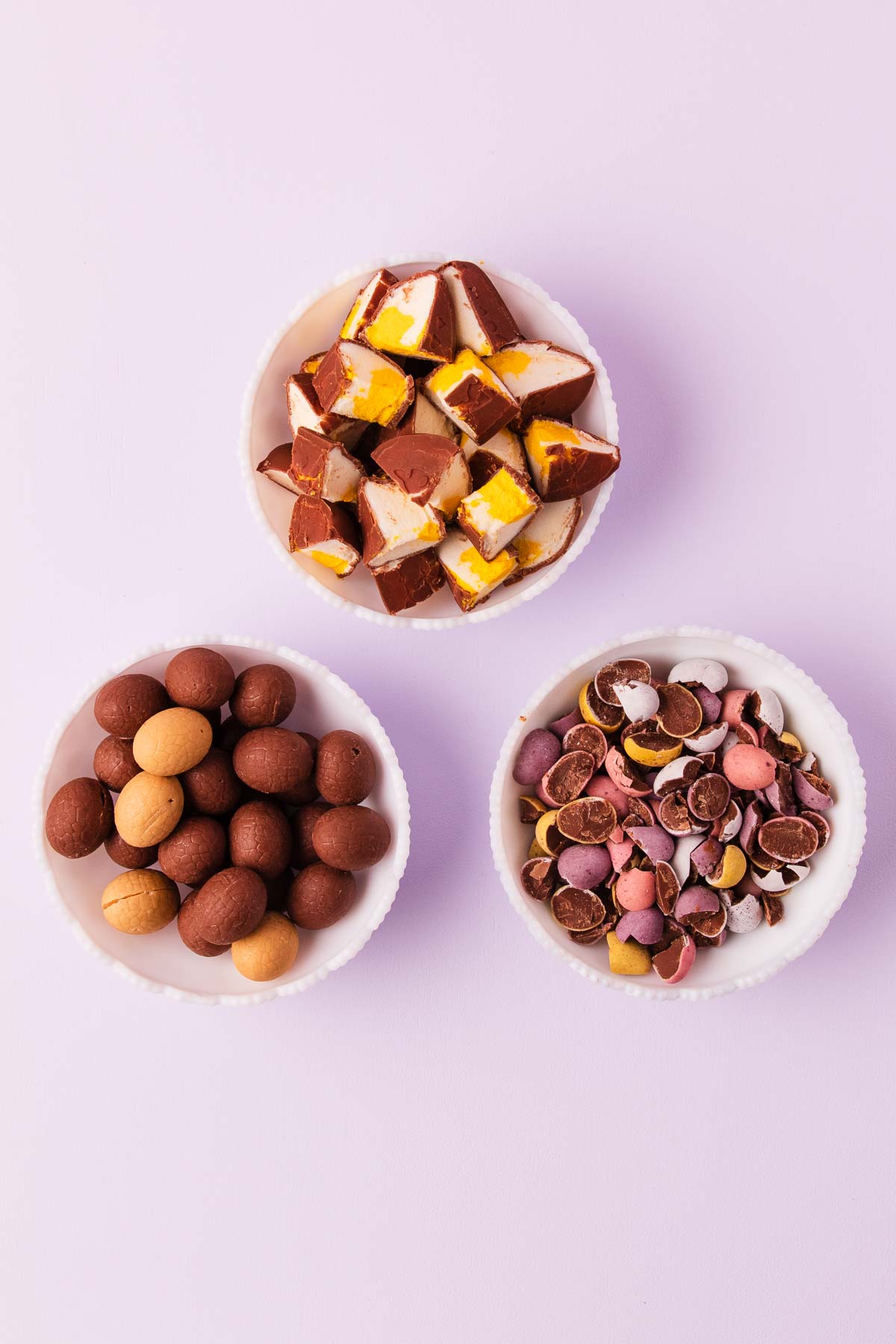 Three white bowls on a light purple background. One bowl has chopped marshmallow easter eggs, one has chopped candy-coated mini eggs and one has small whole plain chocolate eggs.