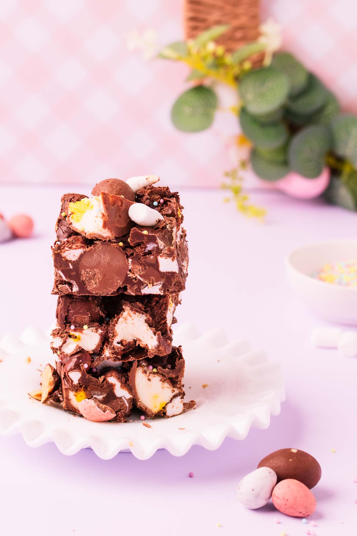 Three stacked pieces of rocky road on a white milk glass plate, with an Easter wreath behind it on a light purple background.