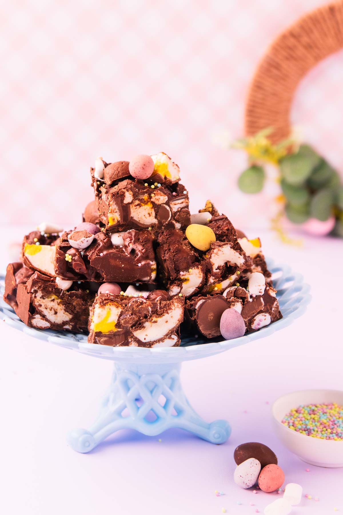 Pieces of Easter egg rocky road piled on a blue basketweave-patterned milk glass cake stand, against a pink and white checkered background, with mini eggs and sprinkles scattered around.