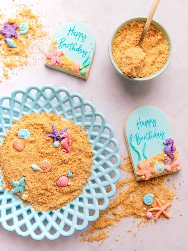 Edible sand on a blue plate, with scattered gumpaste seashells, and two arch-shaped cookies covered with blue fondant and decorated with the sand and seashells.