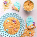Edible sand on a blue milk glass plate, with scattered gumpaste seashells, and two arch-shaped cookies covered with blue fondant and decorated with the sand and seashells.