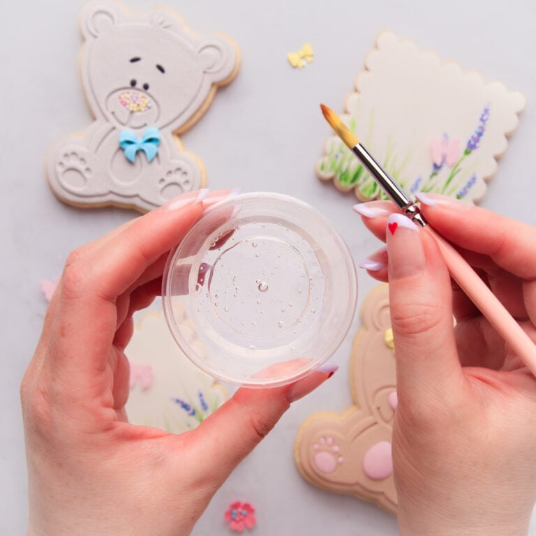 Two hands, holding a small clear container of edible glue in one and a paintbrush in the other, with fondant-covered cookies in the background.