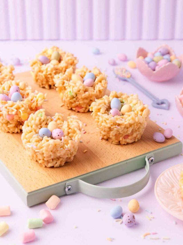 Six Easter rice krispie treat nests on a small wooden board with mint-green painted edges and handle, on a pastel purple background with mini marshmallows, pastel sprinkles and mini eggs scattered around.