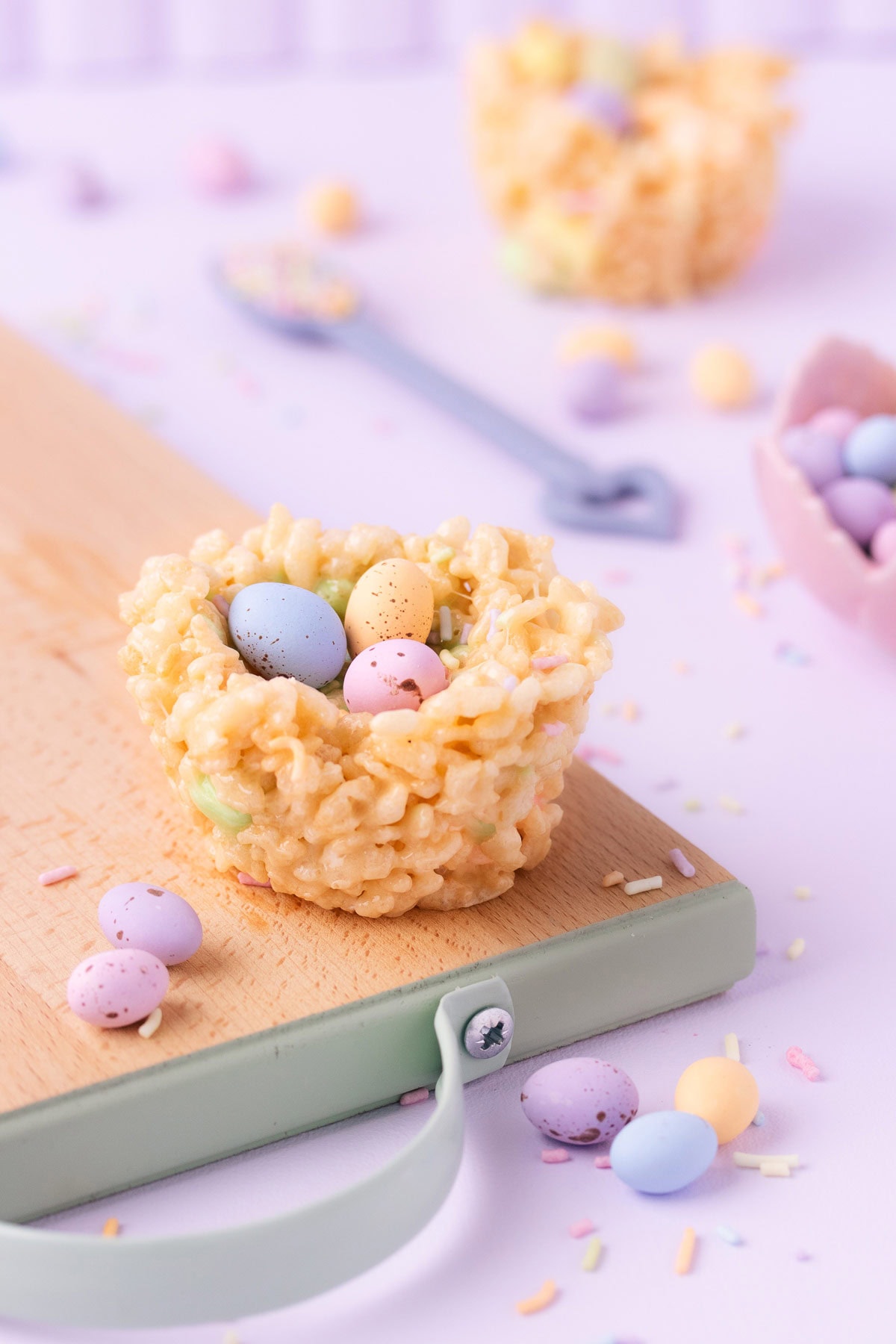 A rice krispie treat nest filled with three pastel mini eggs, on a wooden board with a mint green edge, on a pastel purple background.