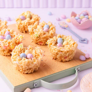 Six Easter rice krispie treat nests on a small wooden board with mint-green painted edges and handle, on a pastel purple background with mini marshmallows, pastel sprinkles and mini eggs scattered around.