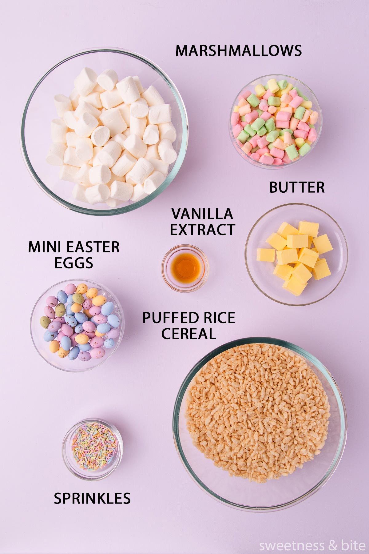 The ingredients - marshmallows, butter, vanilla, sprinkles and mini easter eggs, in bowls on a pastel pink background.