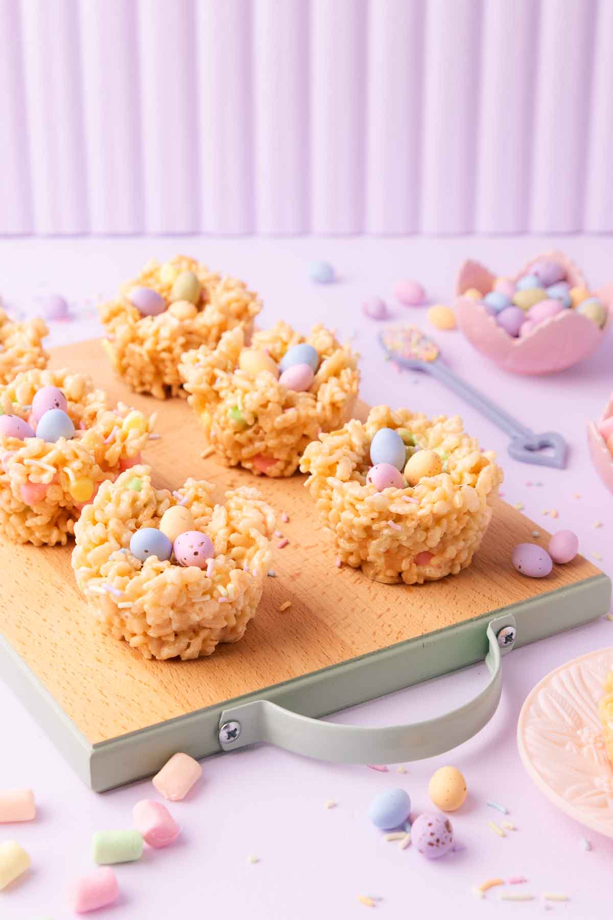 Six Easter rice krispie treat nests on a small wooden board with mint-green painted edges and handle, on a pastel purple background with mini marshmallows, pastel sprinkles and mini eggs scattered around.