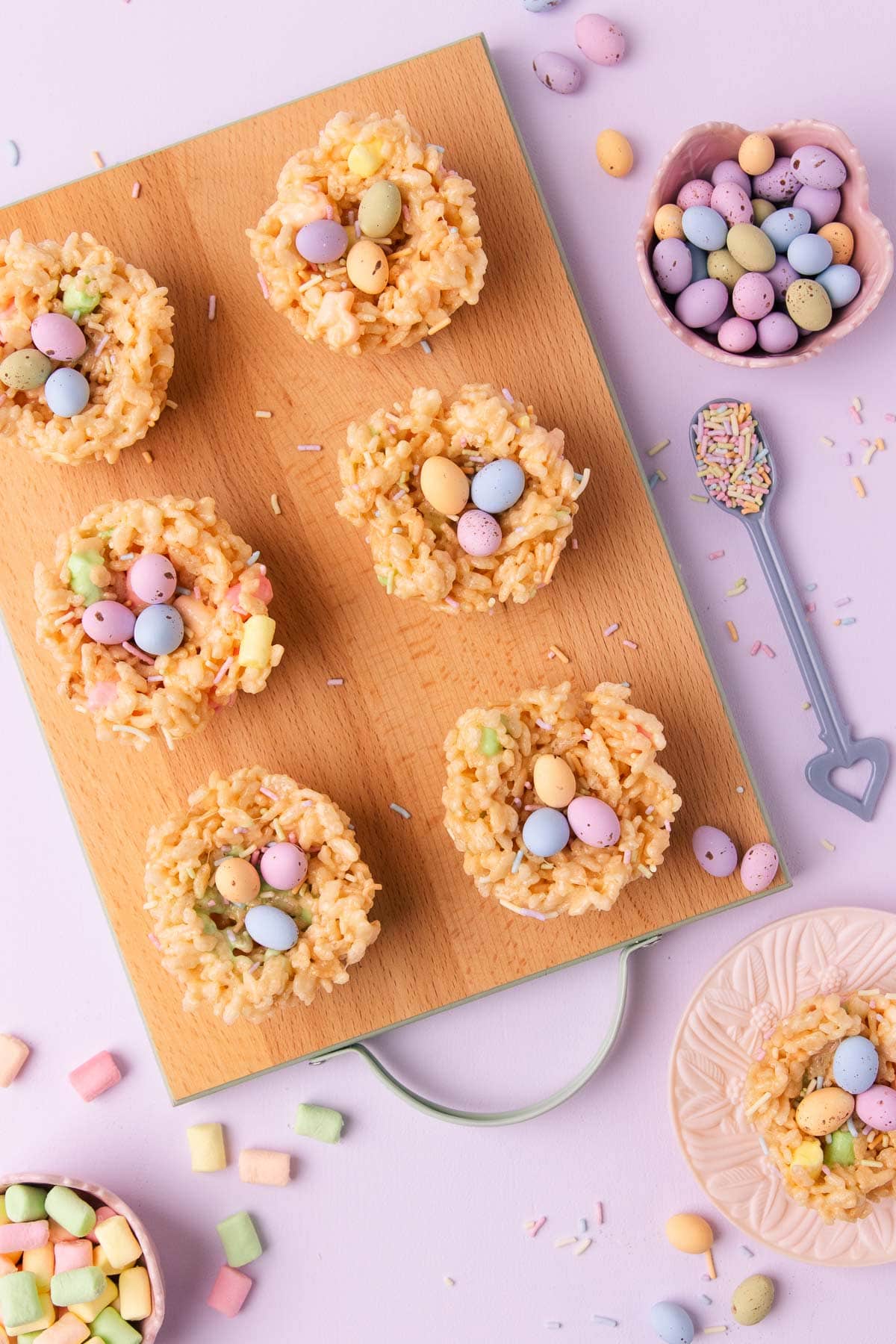 Overhead image of six rice krispie treat nests on a small wooden board with mint-green painted edges and handle, with a small bowl of pastel mini eggs, and small bowl of pastel mini marshmallows and a little blue-handles ceramic spoons filled with pastel sprinkles.