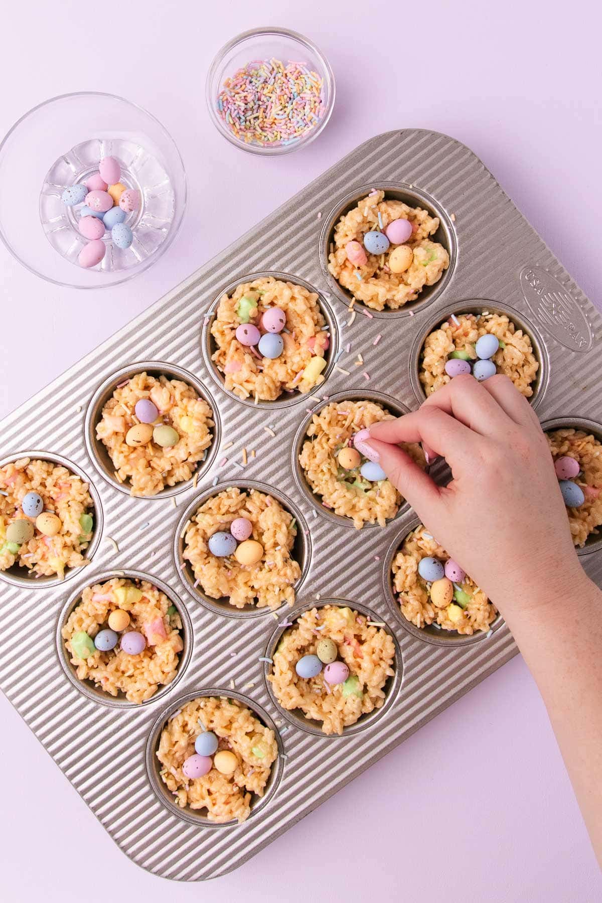 A hand placing mini pastel-coloured candy-coated Easter eggs into the middle of each rice krispie nest.