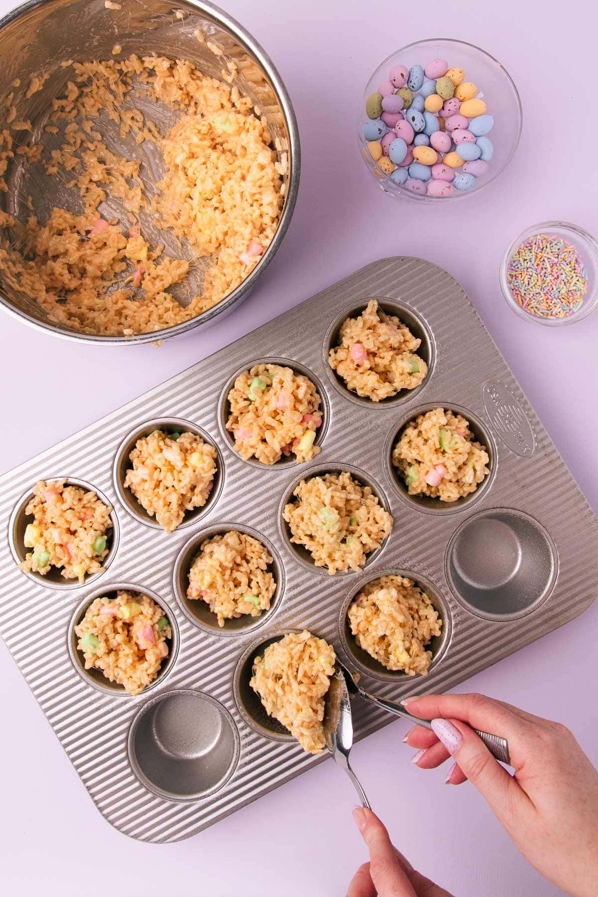 The rice krispie mixture being spooned into the holes of a 12-hole muffin pan.
