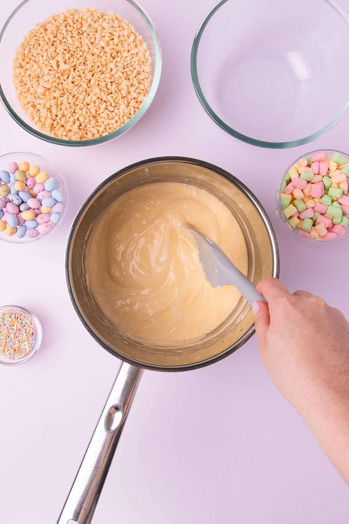A hand stirring the melted marshmallow, butter and vanilla mixture in a large stainless steel saucepan, on a light purple background.