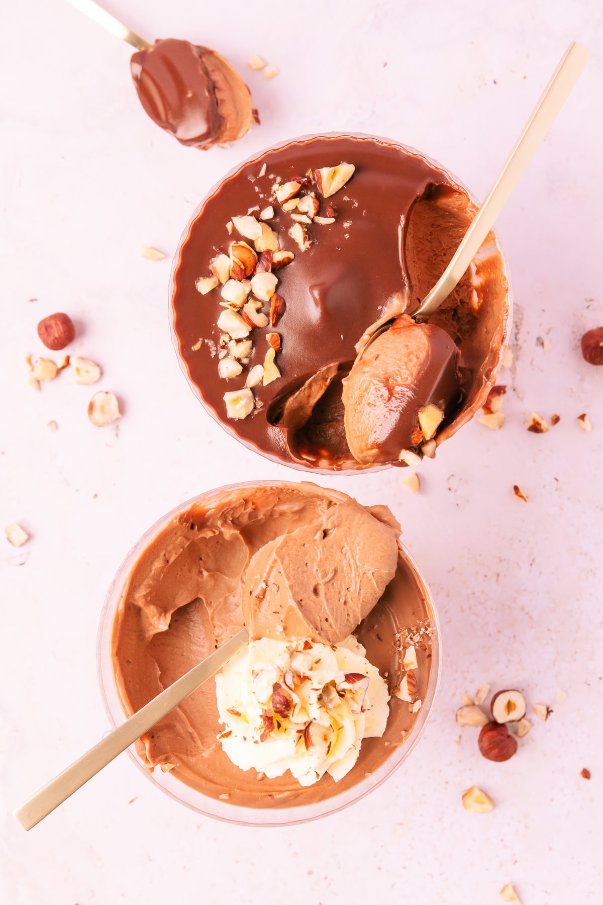 Overhead image of two bowls of mousse, one topped with ganache and one topped with whipped cream, both sprinkled with hazelnuts each with a spoonful of mousse scooped out.