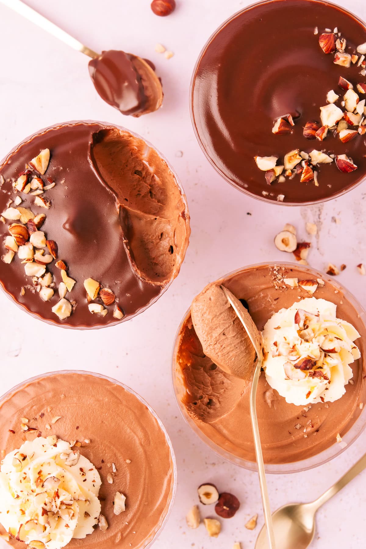 Overhead image of four bowls of mousse, two topped with ganache and two topped with whipped cream, all sprinkled with hazelnuts and two bowls have spoonfuls of mousse scooped out, showing the creamy texture of the mousse.