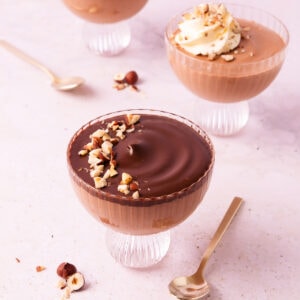 Three scalloped glass footed bowls with Nutella mousse, two are topped with ganache, and one is topped with a swirl of whipped cream.