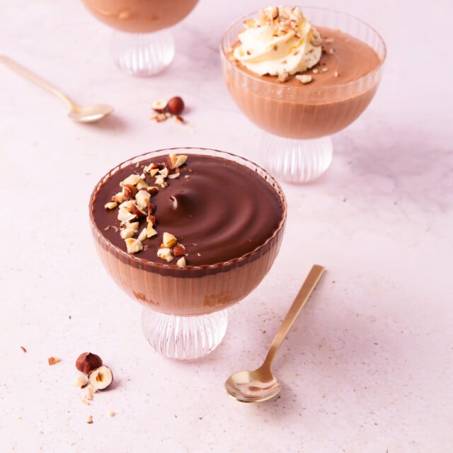 Three scalloped glass footed bowls with Nutella mousse, two are topped with ganache, and one is topped with a swirl of whipped cream.