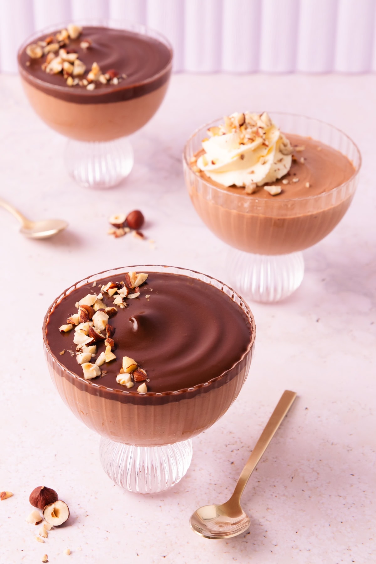 Three scalloped glass footed bowls with Nutella mousse, two are topped with ganache, and one is topped with a swirl of whipped cream.