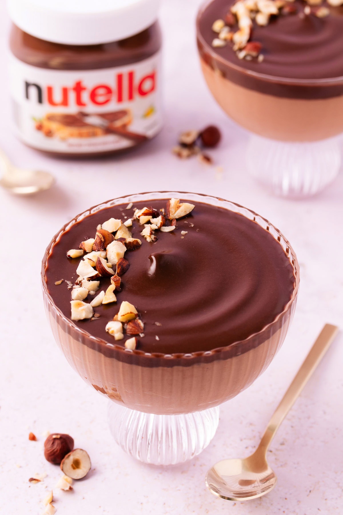 A scalloped glass footed dessert bowl with Nutella mousse and a layer of ganache, sprinkled with chopped hazelnuts on a pinky-purple marble background, with a jar of Nutella.