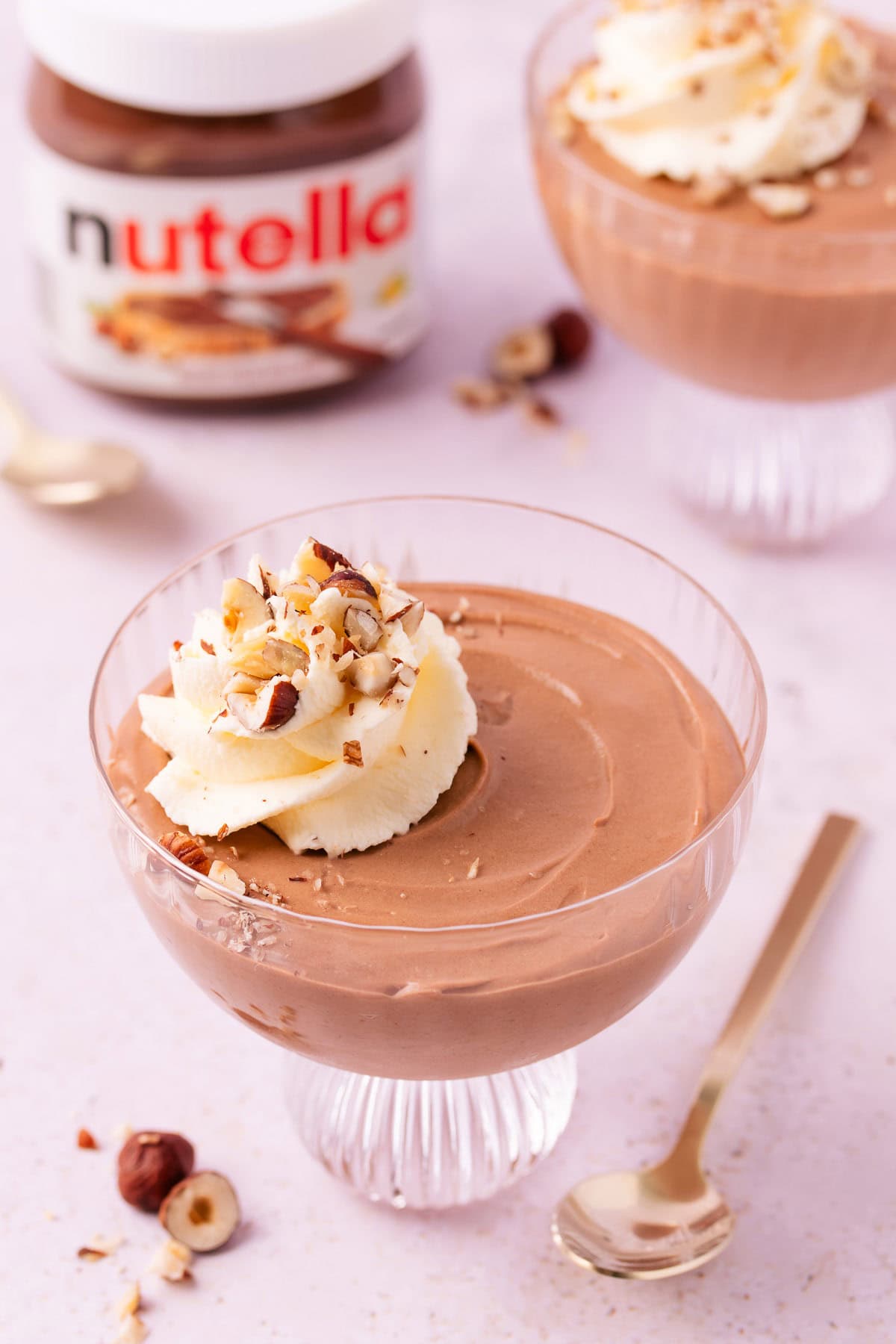 A scalloped glass footed dessert bowl with Nutella mousse and a swirl of whipped cream, sprinkled with chopped hazelnuts on a pinky-purple marble background, with a jar of Nutella.
