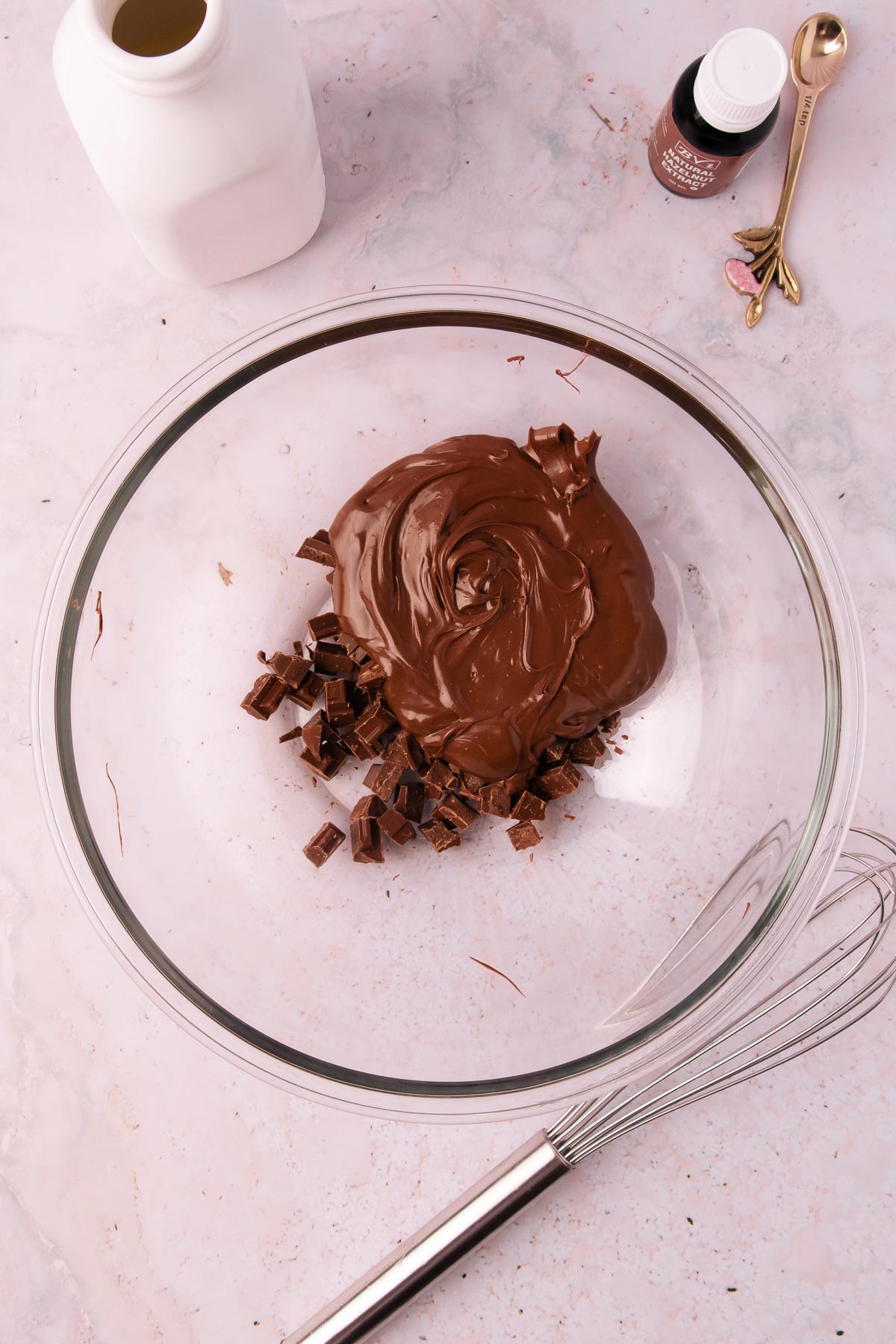 Chopped milk chocolate and Nutella in a large glass bowl, with a whisk and a bottle of hazelnut extract.