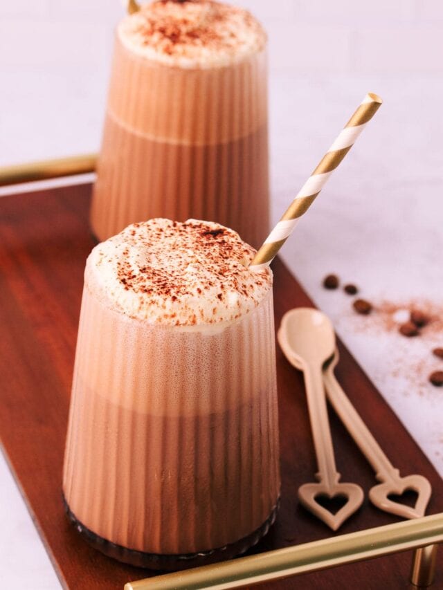 Two mocha milkshakes in glasses with a ribbed texture, topped with whipped cream and a sprinkle of cocoa, with gold and white striped paper straws on a dark wooden board with gold handles.