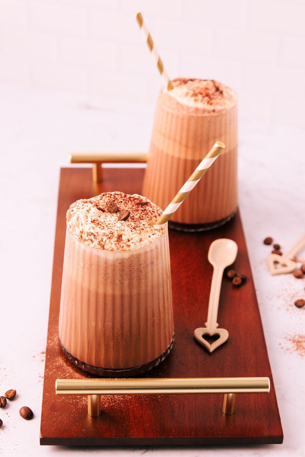 Two mocha milkshakes in glasses with a ribbed texture, topped with whipped cream and a sprinkle of cocoa, with gold and white striped paper straws on a dark wooden board with gold handles.