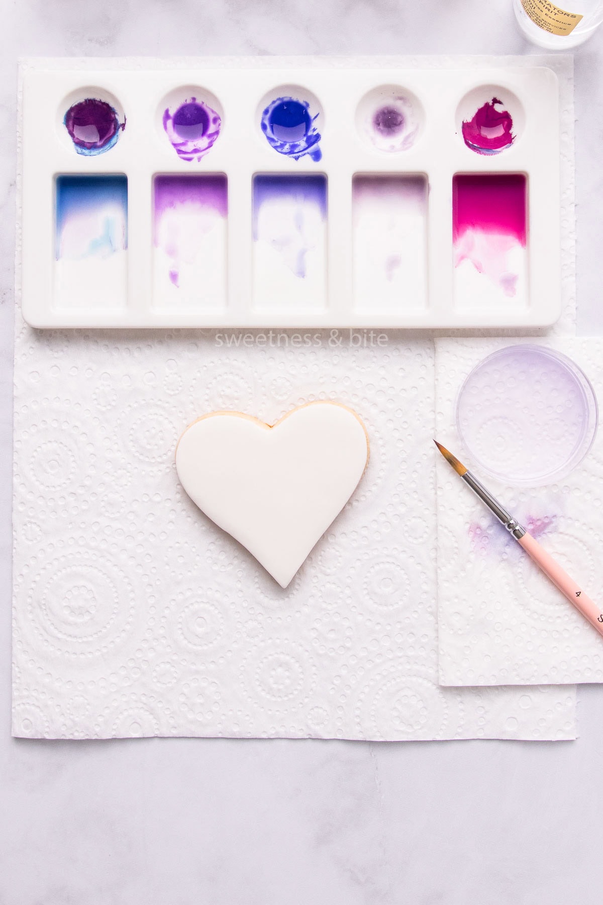 A heart-shaped cookie covered in white fondant icing, with a palette of watercolour paint and a paintbrush.