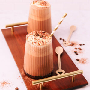 Two mocha milkshakes in glasses with a ribbed texture, topped with whipped cream and a sprinkle of cocoa, with gold and white striped paper straws on a dark wooden board with gold handles.