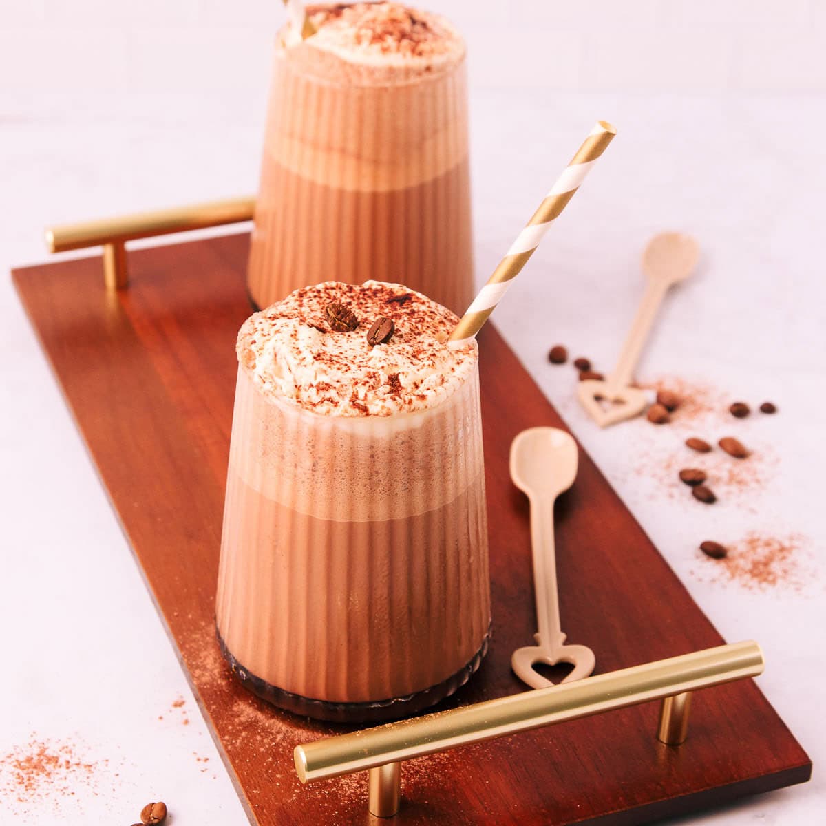 Two mocha milkshakes in glasses with a ribbed texture, topped with whipped cream and a sprinkle of cocoa, with gold and white striped paper straws on a dark wooden board with gold handles.