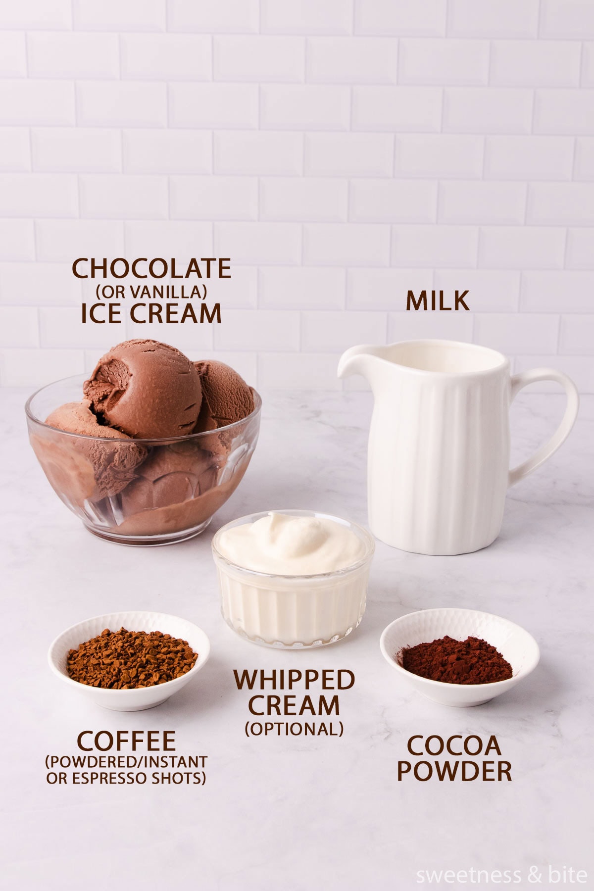 The mocha milkshake ingredients - chocolate ice cream in a glass bowl, milk in a white jug, coffee, cocoa powder and whipped cream in small bowls, on a pale grey marble background.